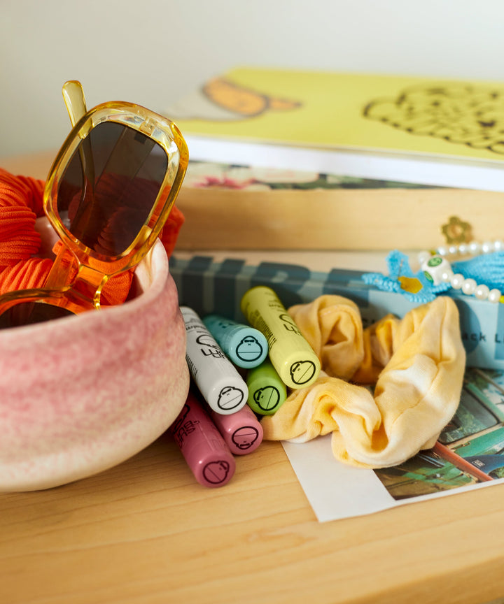 CocoBalm Lip Balm - Wild Strawberry by Sun Bum displayed with colorful accessories including sunglasses, scrunchies, and books.