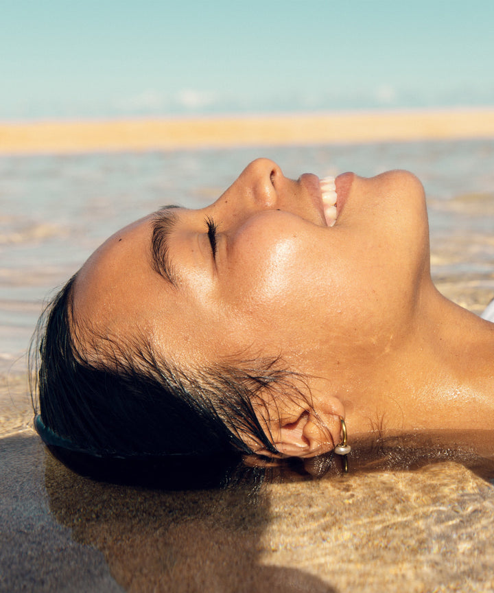Woman smiling while lying on the beach and letting the ocean waves gently flow over her, showcasing the moisturizing effect of Aloe and Vitamin E from the Original SPF 30 Sunscreen Lip Balm - Kiwi by Sun Bum.