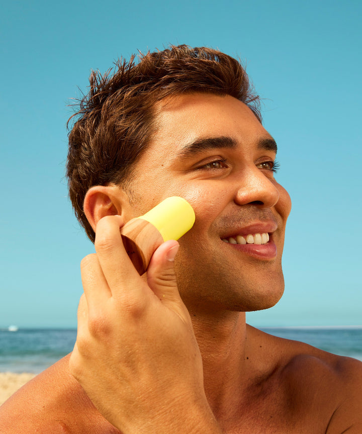 Smiling person applying yellow sunscreen stick to cheek at beach, demonstrating easy application of SPF 40 protection for face and body. Clear blue sky and ocean in background. Sun Bum