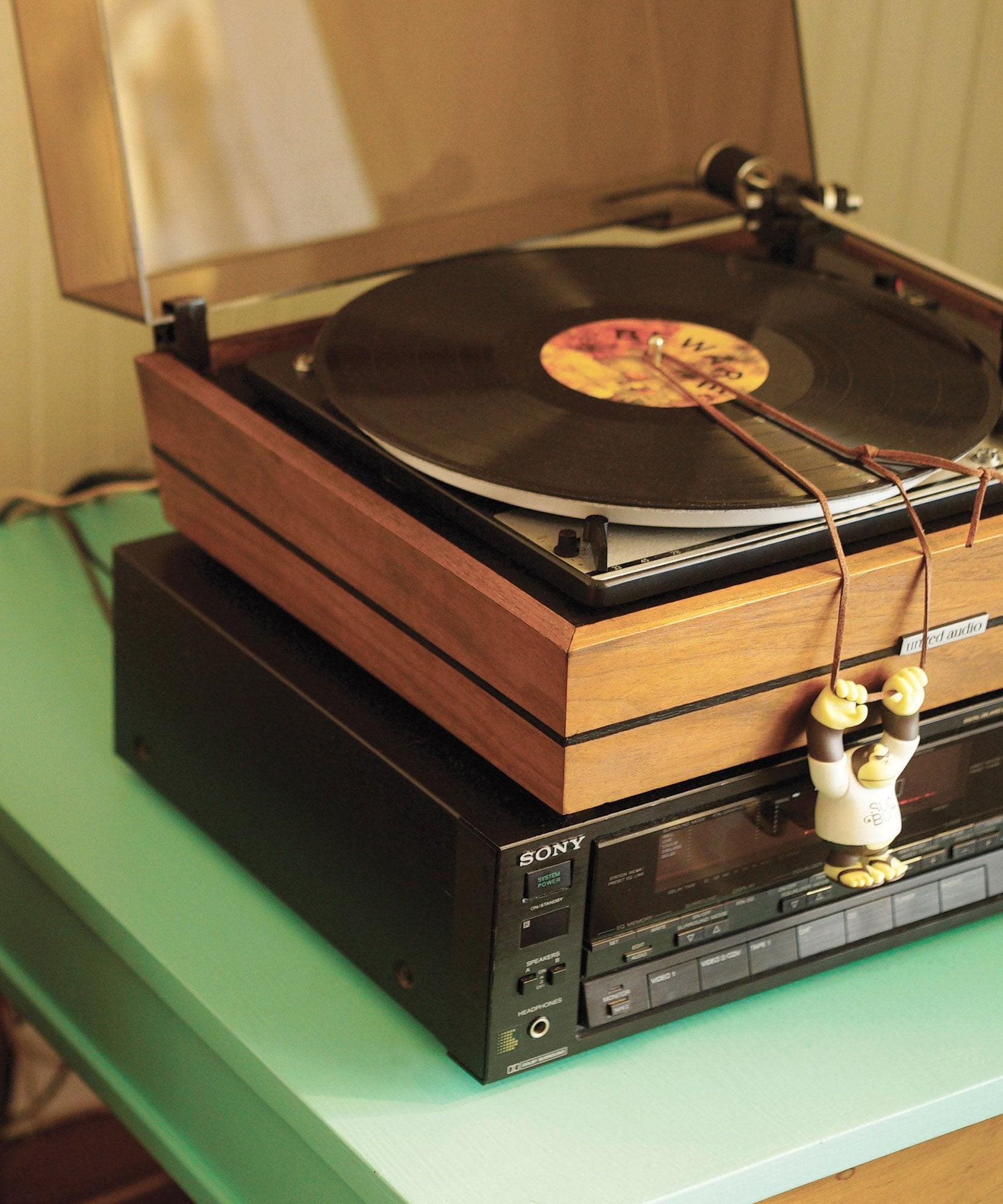 Close-up of 'Lucky Bum' figure hanging from a retro turntable setup, enjoying some good vinyl vibes. Sun Bum.