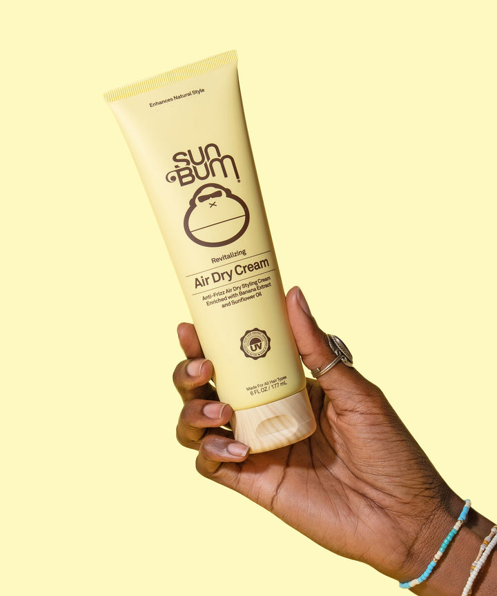 Hand holding a tube of Revitalizing Air Dry Cream by Sun Bum, designed to smooth flyaways and enhance natural hair texture with Banana and Sunflower Oil.