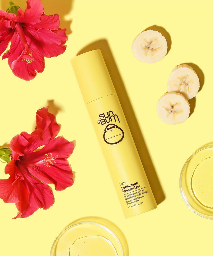 Daily Sunscreen Moisturizer SPF 30 by Sun Bum with antioxidant-rich Banana, Wild Rosella, and Niacinamide, surrounded by red hibiscus flowers and banana slices on a yellow background.