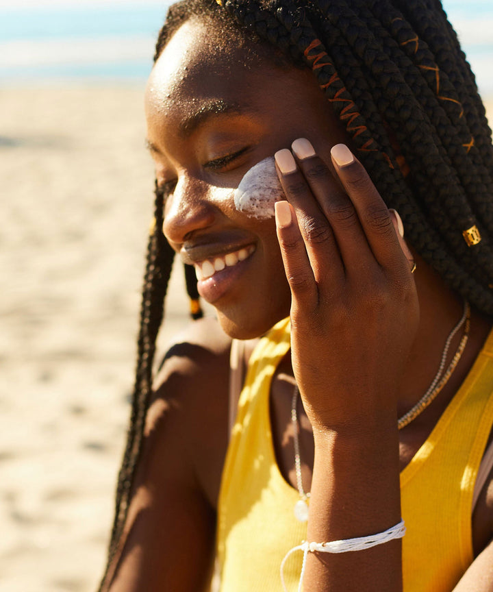 Woman applying Daily Sunscreen Moisturizer SPF 30 at the beach, keeping her skin hydrated and protected with antioxidant-rich Banana, Wild Rosella, and Niacinamide. Sun Bum,