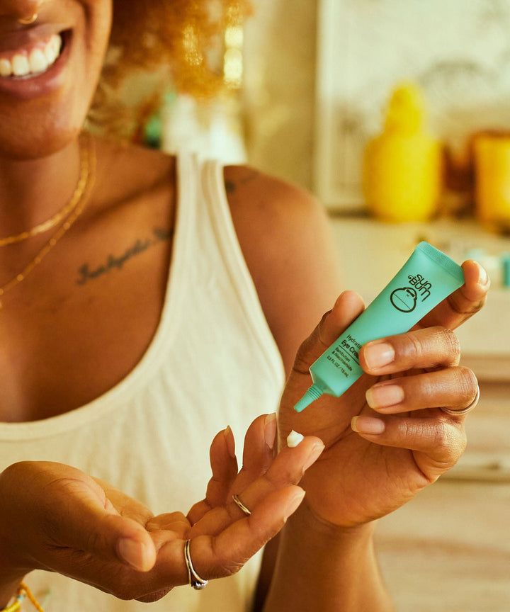 Woman applying hydrating eye cream to hand with Sun Bum product packaging visible in a cozy bathroom setting.