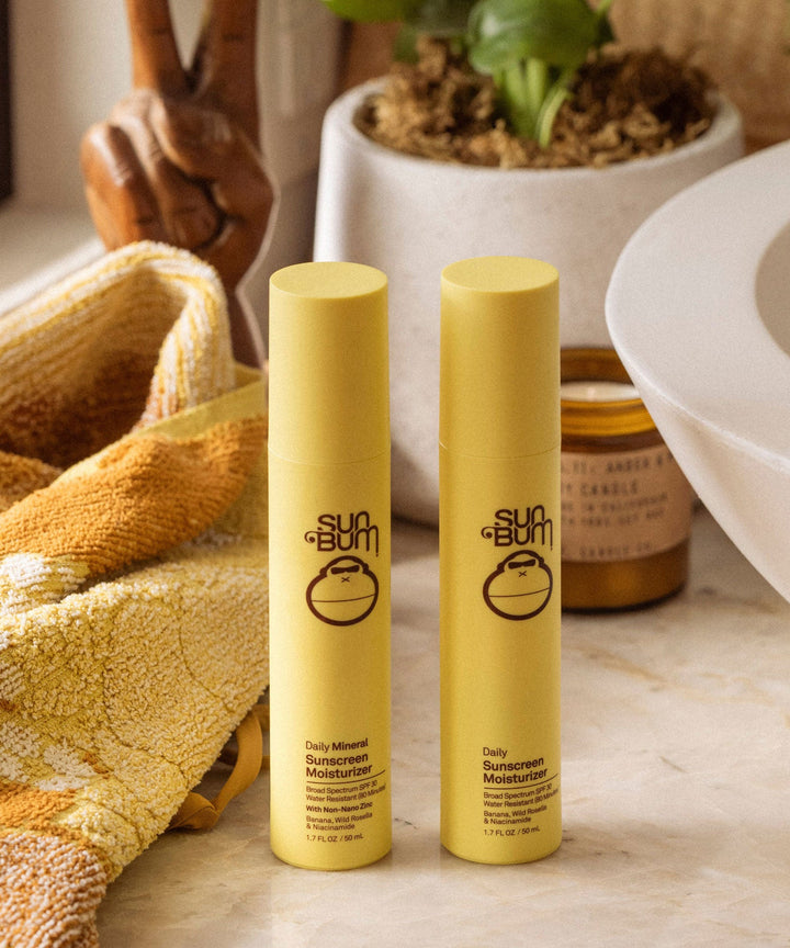Two bottles of Sun Bum Daily Mineral Sunscreen Moisturizer SPF 30 on a bathroom counter with a towel and a candle in the background. Sun Bum