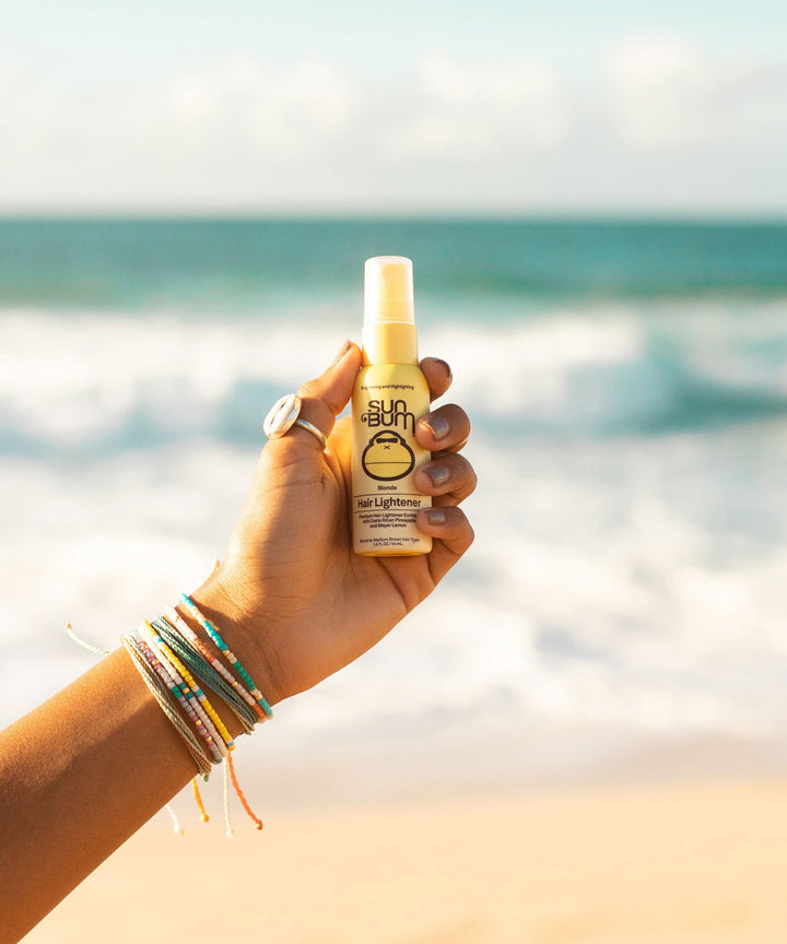 Hand holding a bottle of Blonde Hair Lightener spray by the beach, enhancing hair naturally with Costa Rican Pineapples and Meyer Lemons. Sun Bum.