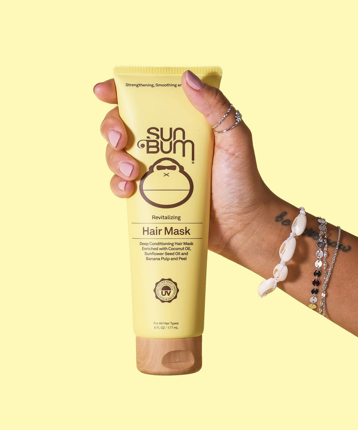 Person holding a tube of Revitalizing Hair Mask by Sun Bum, designed to nourish dry and damaged hair, making it softer and smoother.