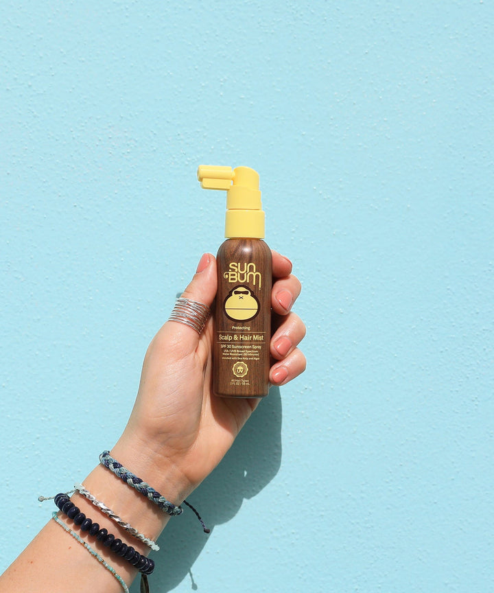 Hand holding Sun Bum Scalp & Hair Mist SPF 30 bottle against a turquoise background, Sun Bum.