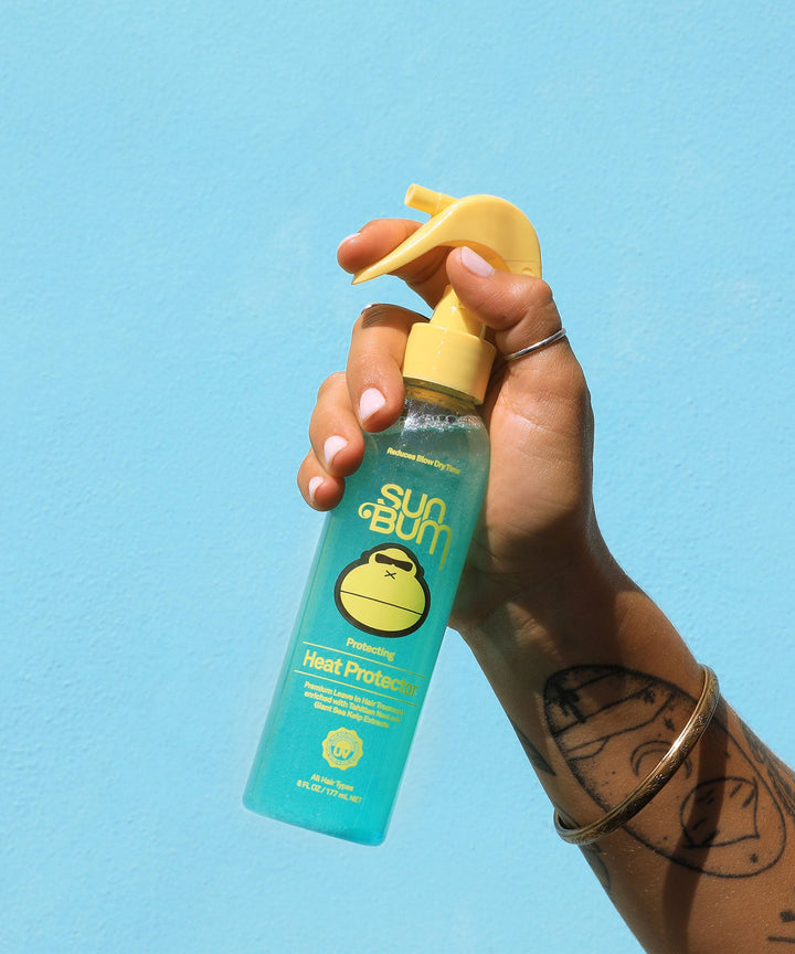 Hand holding a bottle of Sun Bum Heat Protector spray against a bright blue background, designed to reduce blow-dry time and shield hair from frizz and thermal heat damage, Sun Bum