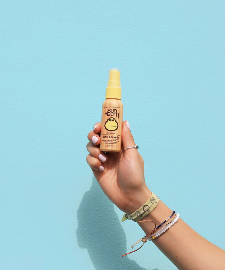 Hand holding a bottle of Sun Bum 3 In 1 Leave In hair care spray against a light blue background.