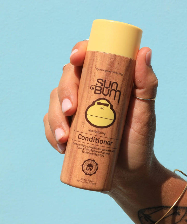 Hand holding a bottle of Revitalizing Conditioner by Sun Bum, featuring hydrating ingredients like Coconut Oil, Banana, and Sunflower Seed Oil.