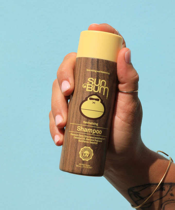 Hand holding a bottle of Sun Bum Revitalizing Shampoo with Coconut Oil, Banana, and Sunflower Seed Oil against a light blue background, Sun Bum.
