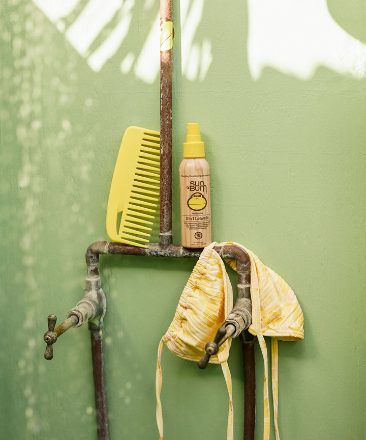 Yellow comb, 3 In 1 Leave In hair treatment bottle, and striped yellow headscarf on rustic shower fixtures against light green background - Sun Bum.