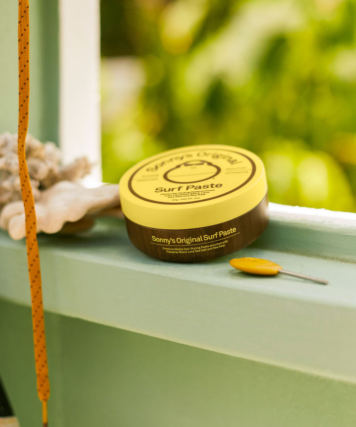 Texturizing Surf Paste in a yellow container sits on a windowsill with greenery in the background. Ideal for short hair styling with ease. Sun Bum.