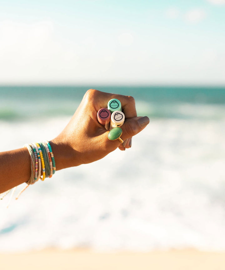 Hand holding three CocoBalm Lip Balms in a 3-pack with ocean in the background, Sun Bum.