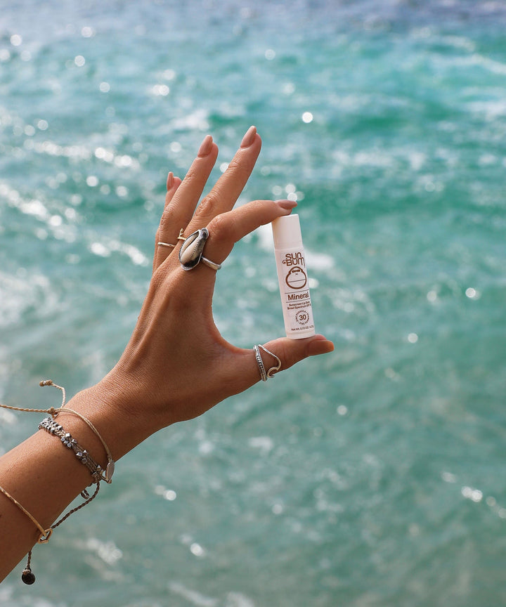 Mineral SPF 30 Sunscreen Lip Balm held by a hand with ocean background, fragrance-free zinc-based protection for your lips. Sun Bum.