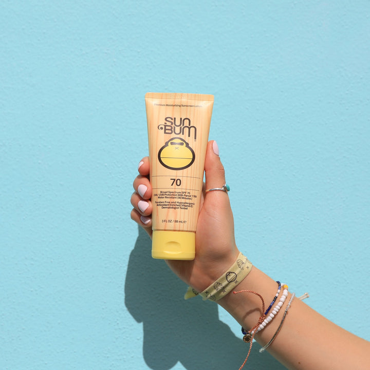 Hand holding a tube of Original SPF 70 Sunscreen Lotion against a blue background, Sun Bum