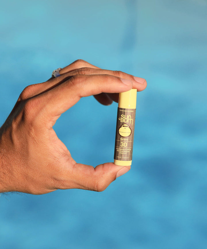 Hand holding a tube of Original SPF 30 Sunscreen Lip Balm - Banana flavor with Aloe and Vitamin E, against a blue background, from Sun Bum.