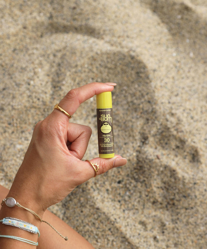 Hand holding the Original SPF 30 Sunscreen Lip Balm in Key Lime flavor with Aloe and Vitamin E by Sun Bum in a beach setting, Sun Bum.