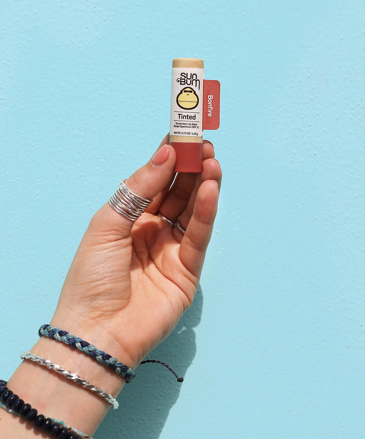 Hand holding Sun Bum tinted SPF 15 lip balm in Bonfire shade with light blue background, Sun Bum.