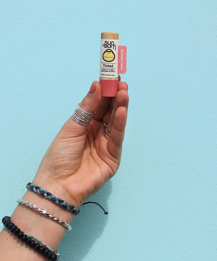 Hand holding a tube of Tinted SPF 15 Lip Balm in shade Sunset Cove against a light turquoise background, Sun Bum.