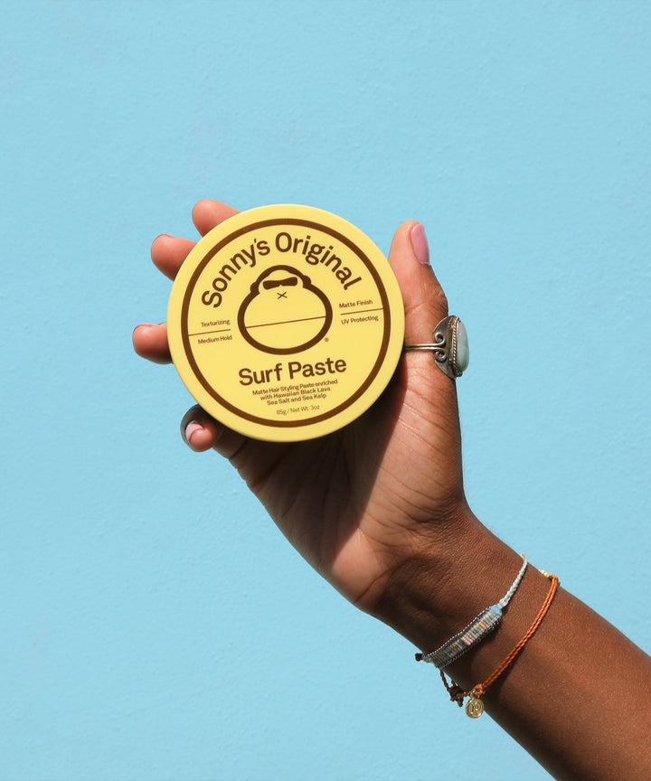 Hand holding a container of Sonny's Original Surf Paste against a blue background, an easy-to-use hairstyling product for short hair, Sun Bum.